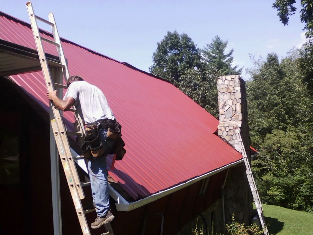 Roofer on a ladder beside a red metal roof and stone chimney — Soffit & Fascia Repair by Roofing Brothers in Mcdaniel